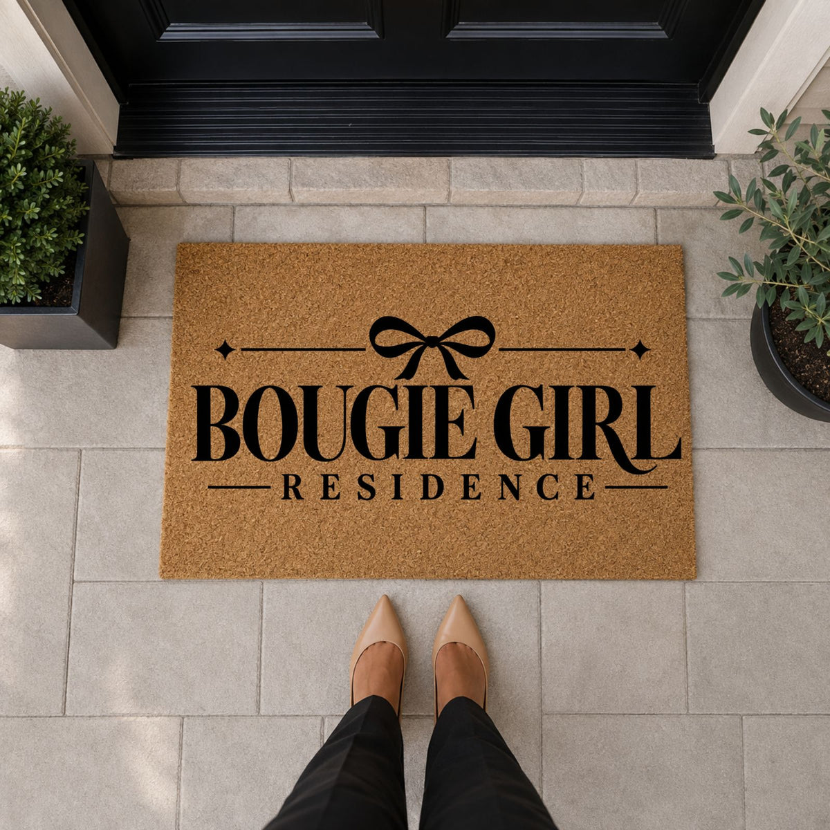 Natural tan Bougie Girl Residence doormat with black lettering, a bow, and sparkle accents in front of a black door.