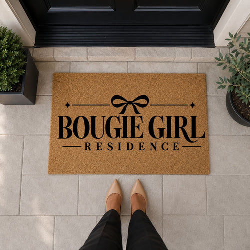 Natural tan Bougie Girl Residence doormat with black lettering, a bow, and sparkle accents in front of a black door.
